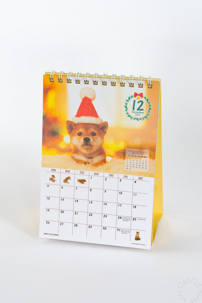 Desk calendar with Shiba Inu in red hat cover, spiral rings, month grid.
