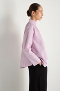 A woman in profile poses against a white backdrop, wearing the Asymmetric Cotton Lilac Shirt with stitched stripes and black pants. The shirt's high collar and her sleek hairstyle add a touch of refined style as she rests one hand in her pocket.