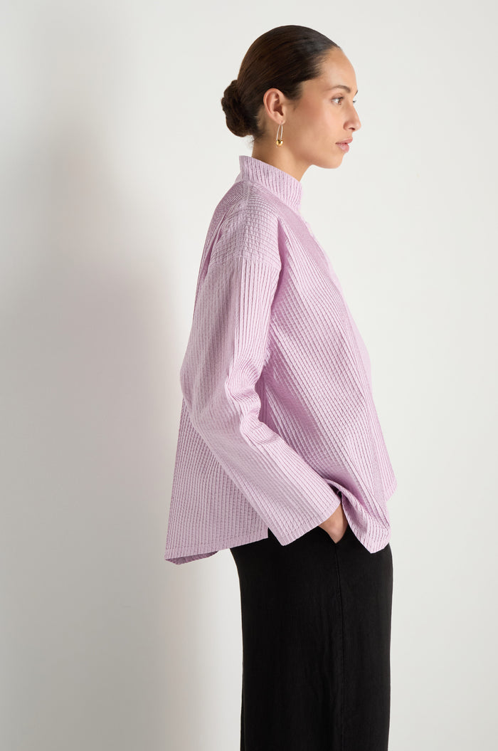 A woman in profile poses against a white backdrop, wearing the Asymmetric Cotton Lilac Shirt with stitched stripes and black pants. The shirt's high collar and her sleek hairstyle add a touch of refined style as she rests one hand in her pocket.