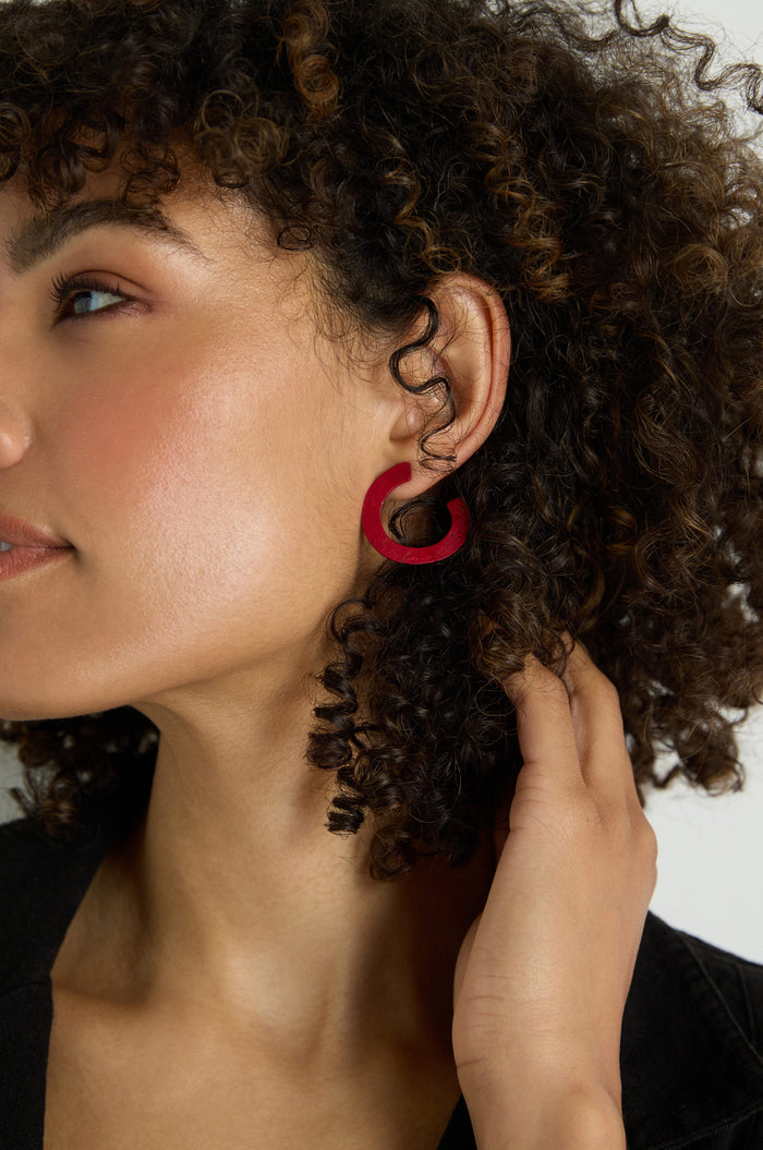 A woman with curly hair, facing slightly away from the camera, wears C Curve Earrings in red anodised aluminium and touches her hair.