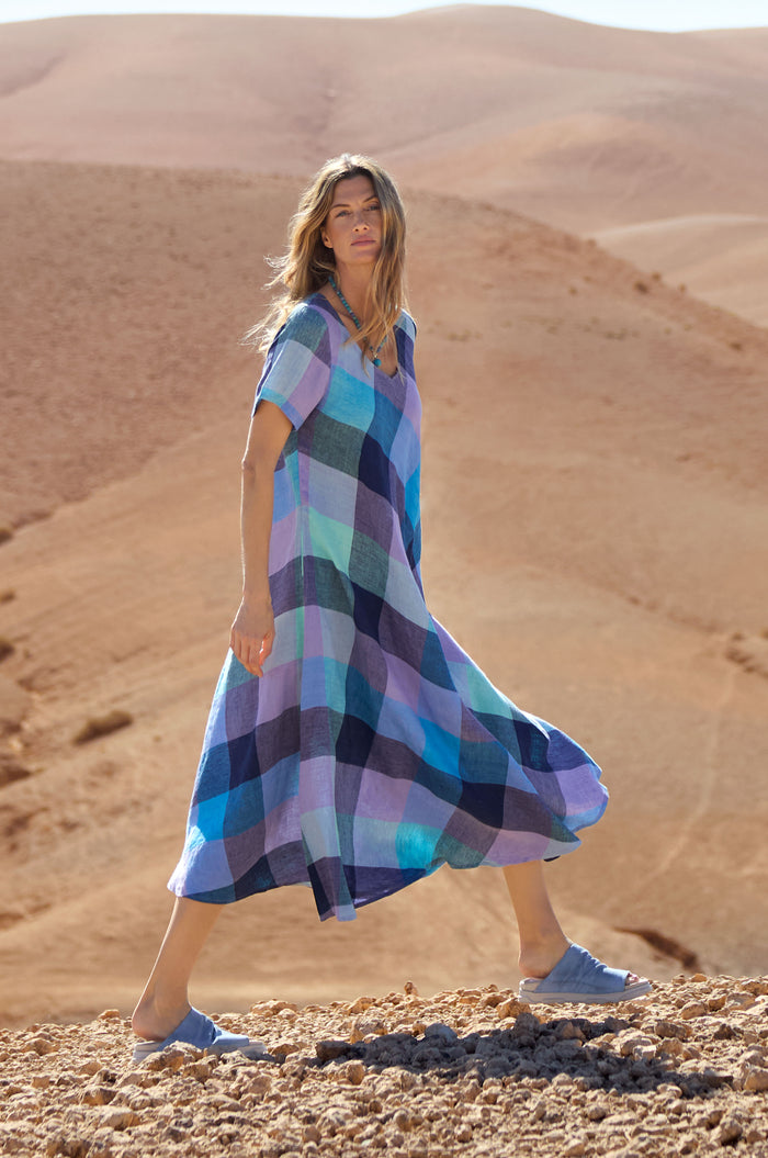 A woman in a loose Cross Dye Check Linen Maxi Dress walks on rocky ground in a desert landscape, with sandy hills in the background.