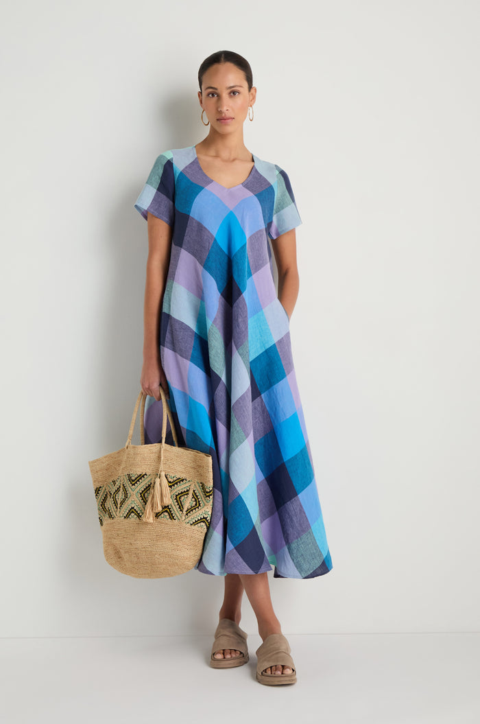 A woman stands against a white wall wearing the Cross Dye Check Linen Maxi Dress in blue and purple plaid, paired with sandals and holding a woven tote bag.
