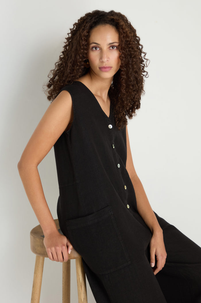 A person with curly hair sits on a wooden stool, wearing the Linen Relaxed Jumpsuit in black with a sleeveless, relaxed fit, facing the camera against a plain light background.