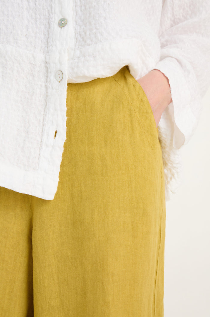 A person wears a textured white button-up and Linen Tulip Hem Golden Bubble Trousers in a relaxed fit, standing with one hand in their pocket.