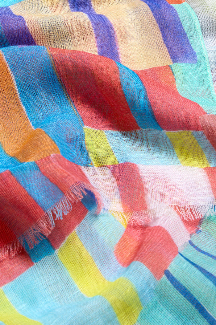 A close-up of the Vivid Painted Stripes Scarf, crafted from cotton and linen, features colorful red, blue, orange, yellow, and green stripes in both directions and a frayed edge.