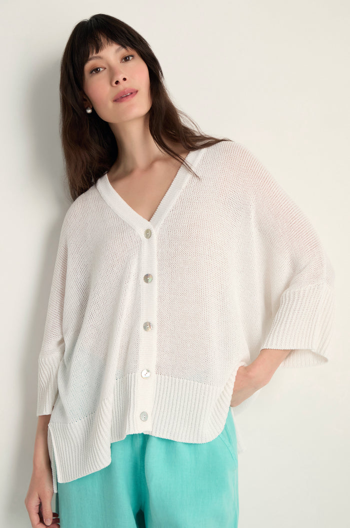 Woman wears the Relaxed White Cardigan—a loose, open-knit white button-up—paired with light blue pants against a plain background. Perfect lightweight layering piece.