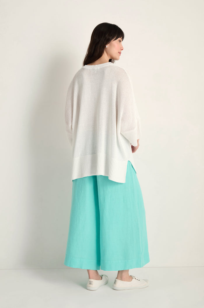 A woman stands against a plain background, facing away. She wears the Relaxed White Cardigan, turquoise wide-leg pants, and white slip-on shoes.