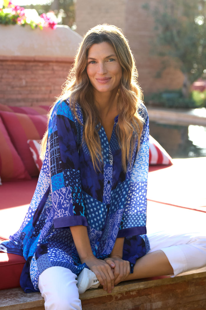 A woman with long, wavy hair sits cross-legged on a red outdoor sofa, smiling at the camera in the Sahara Indigo Patchwork Longline Shirt and white pants.