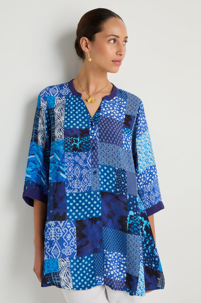 A woman stands against a plain light wall, looking to the side, wearing the Sahara Indigo Patchwork Longline Shirt with white details.