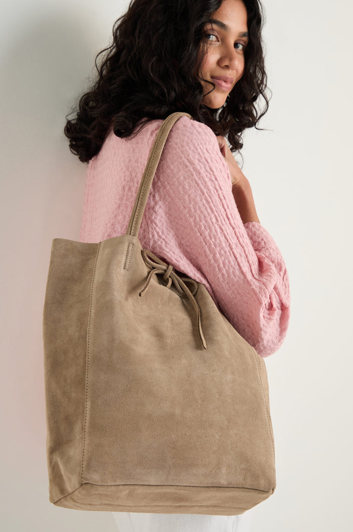 A person with curly hair wears a textured pink top and carries the Soft Suede Sand Tote Bag over their shoulder.