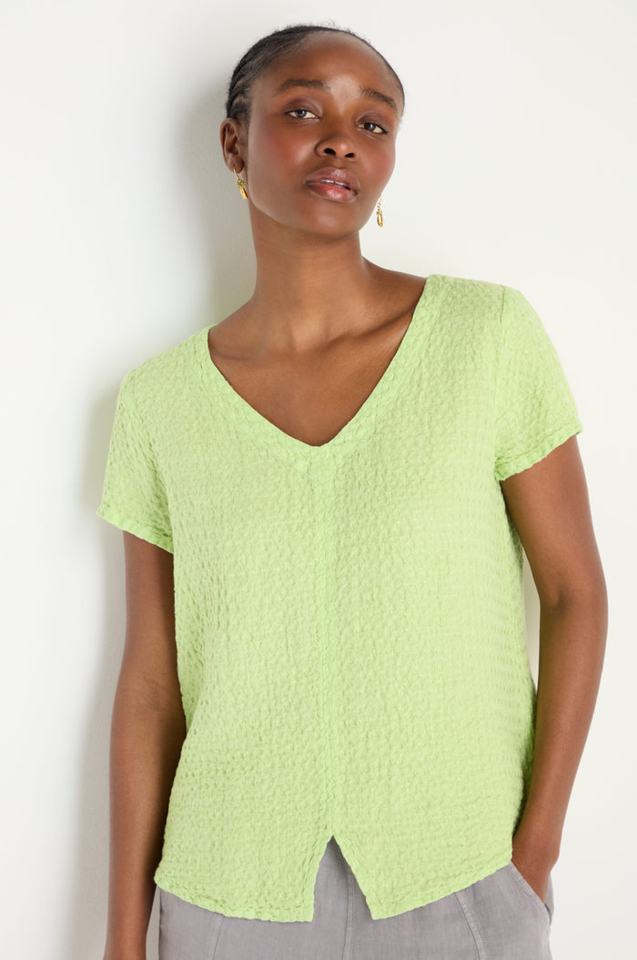 A woman stands against a white wall, wearing the Waffle Linen Lime V Neck Top with short sleeves, paired with gray pants.
